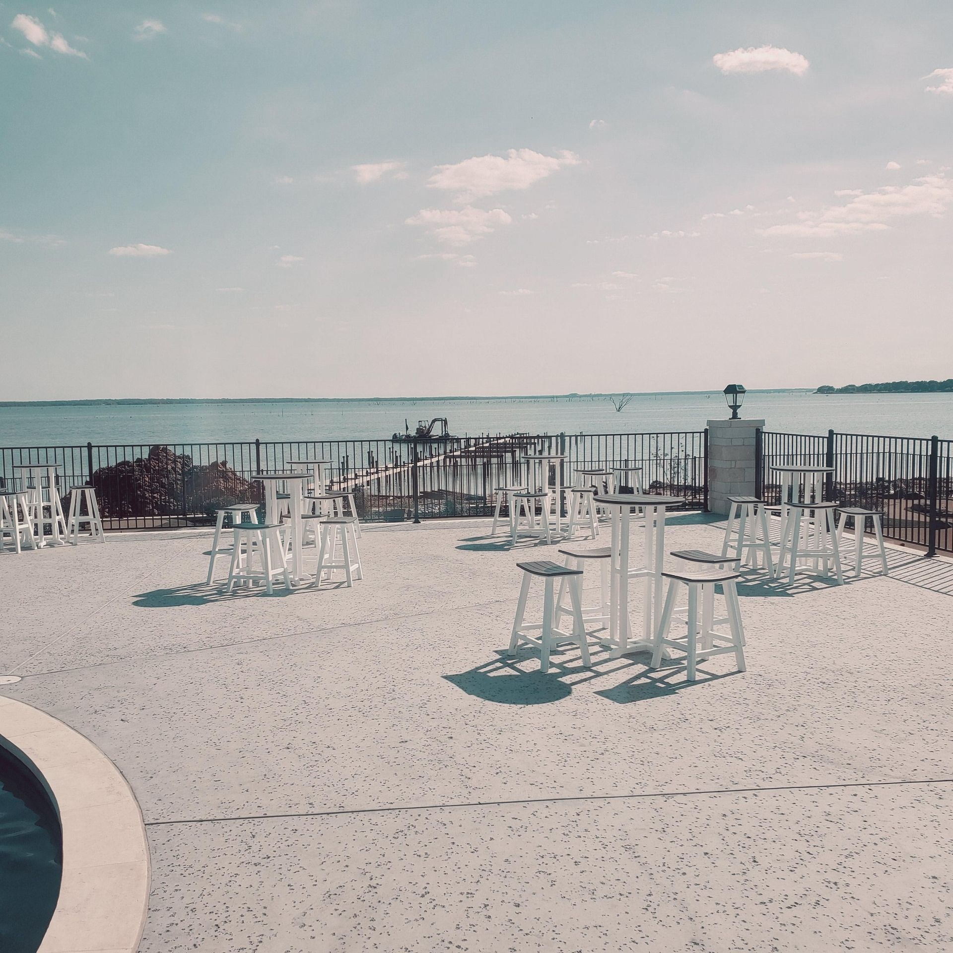 Empty seaside patio with white stools and tables overlooking calm water under a pale sky