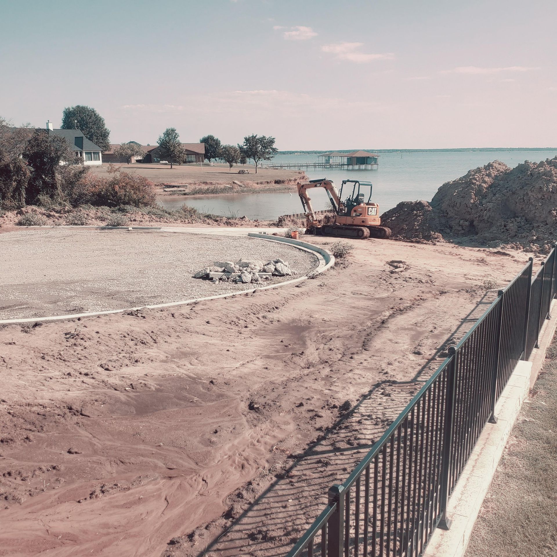 Construction site with excavator beside a waterfront, dirt roads, and a black fence in the foreground
