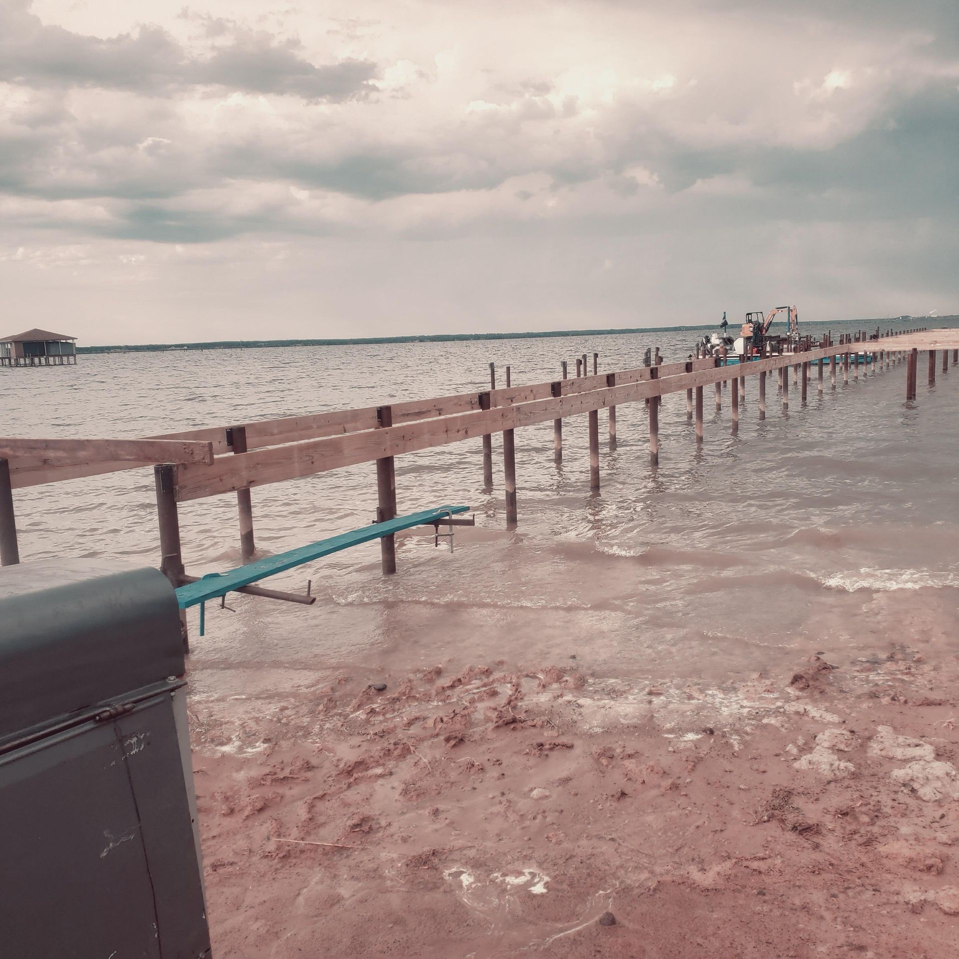 Flooded wooden pier extending into choppy water under a cloudy sky, with a dark railing in the foreground