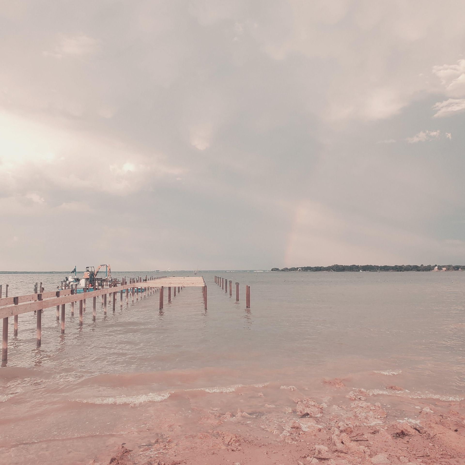 Pink-hued beach with a wooden pier extending into calm water under a cloudy sky