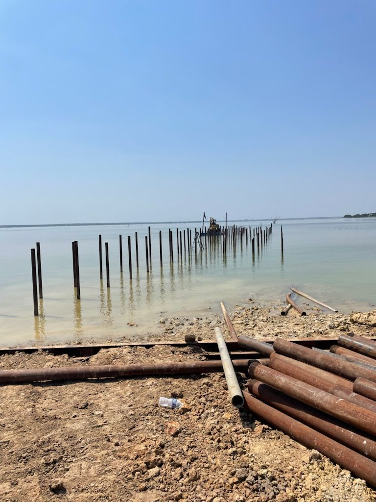Rusty pipes and weathered posts on a pebbled beach beside calm blue water under a clear sky