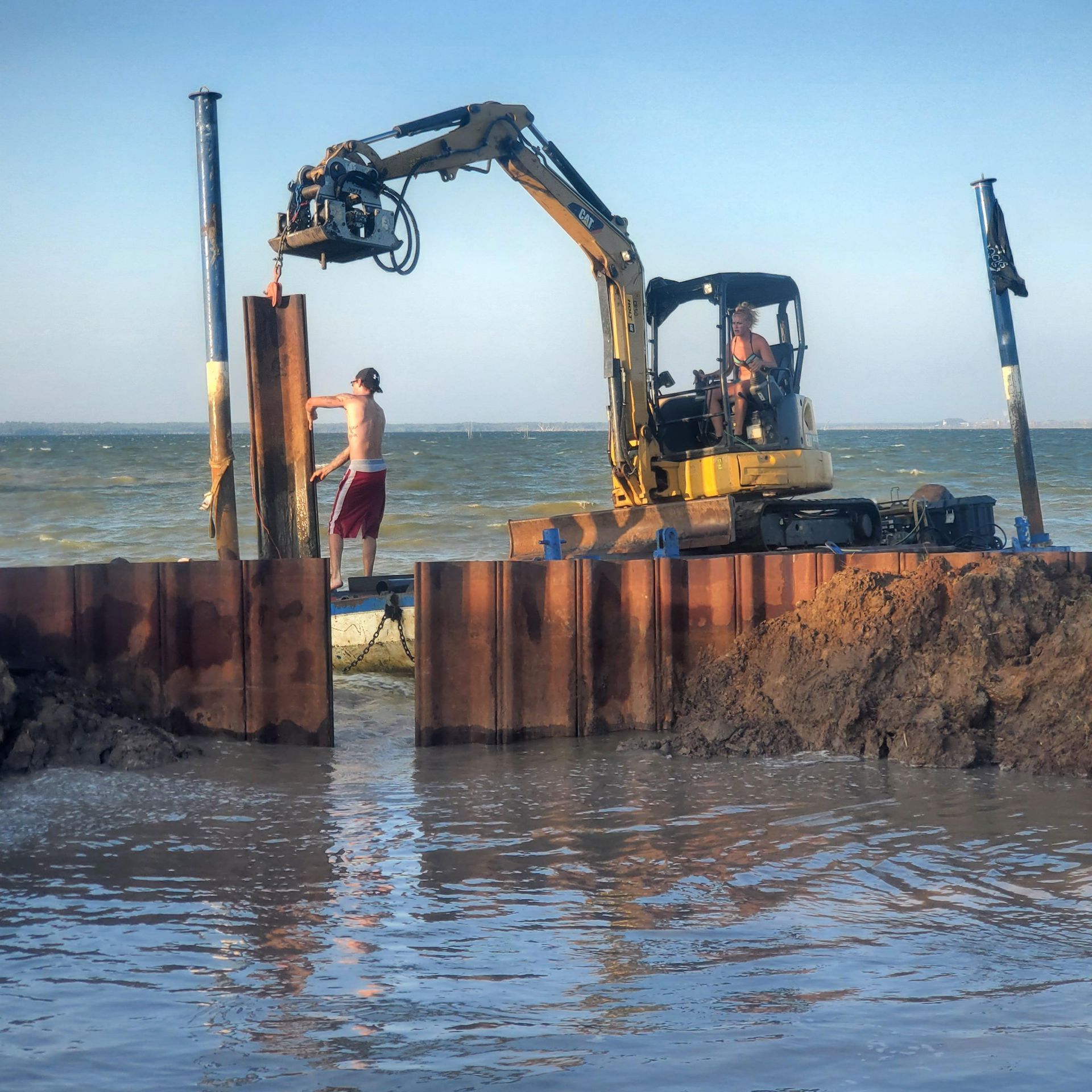 Excavator on a rusted seawall beside the ocean, with a person standing nearby.