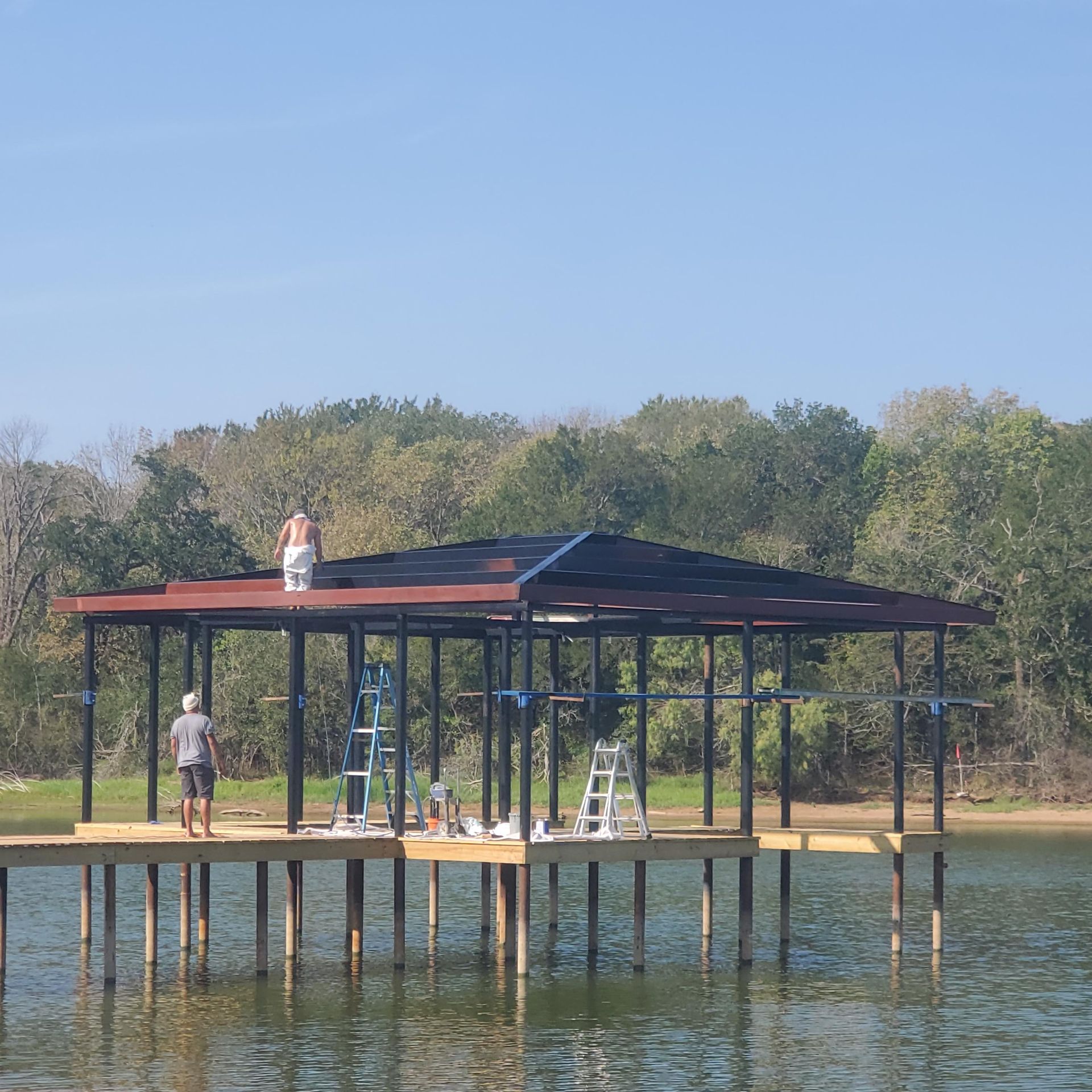 Workers roofing a lakeside pavilion with dark panels and ladders, surrounded by water and trees.