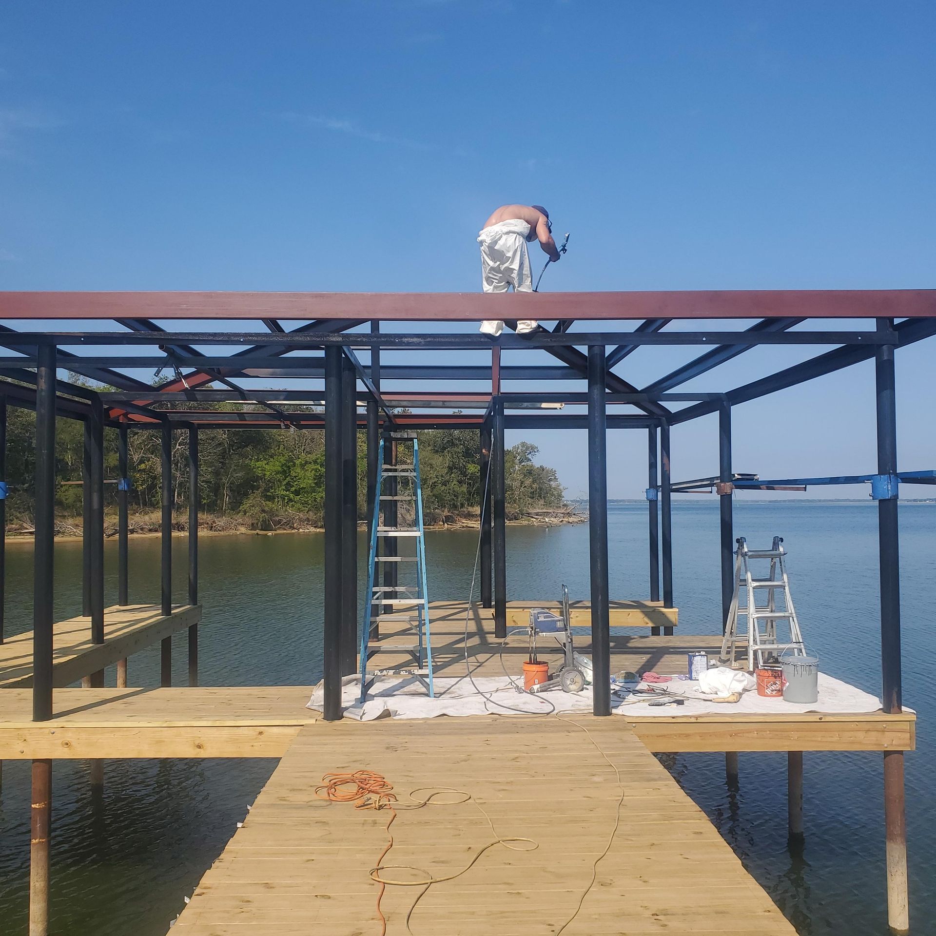 Worker on a dock frame painting the roof, with a ladder and calm water in the background