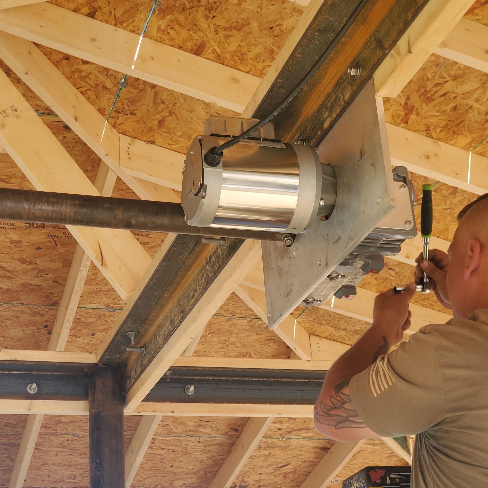 Worker installing a metal motor assembly on exposed ceiling beams in a building frame