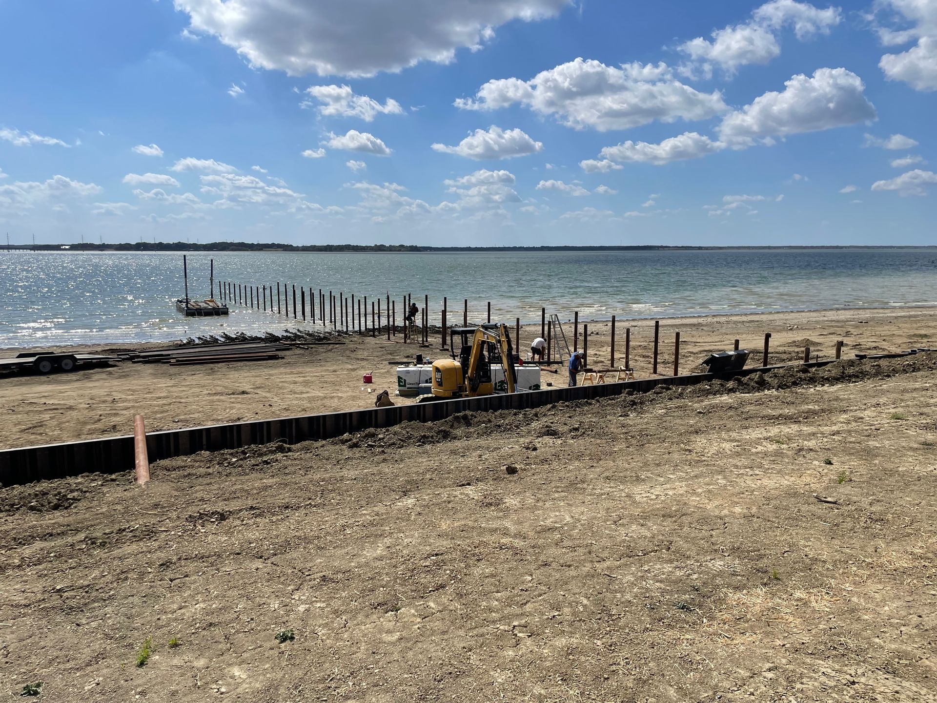 Sandy beach with shoreline markers, construction equipment, and a calm blue lake under a partly cloudy sky
