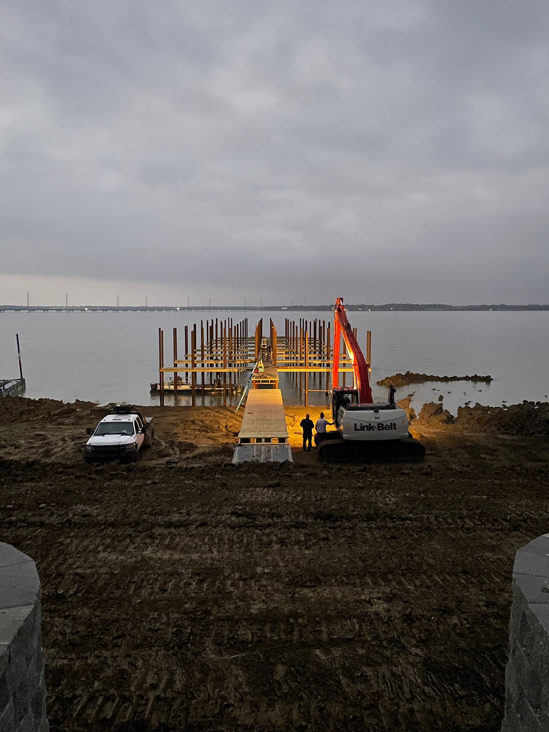Construction barge with orange crane and lights on a calm waterfront at dusk, with two vehicles nearby