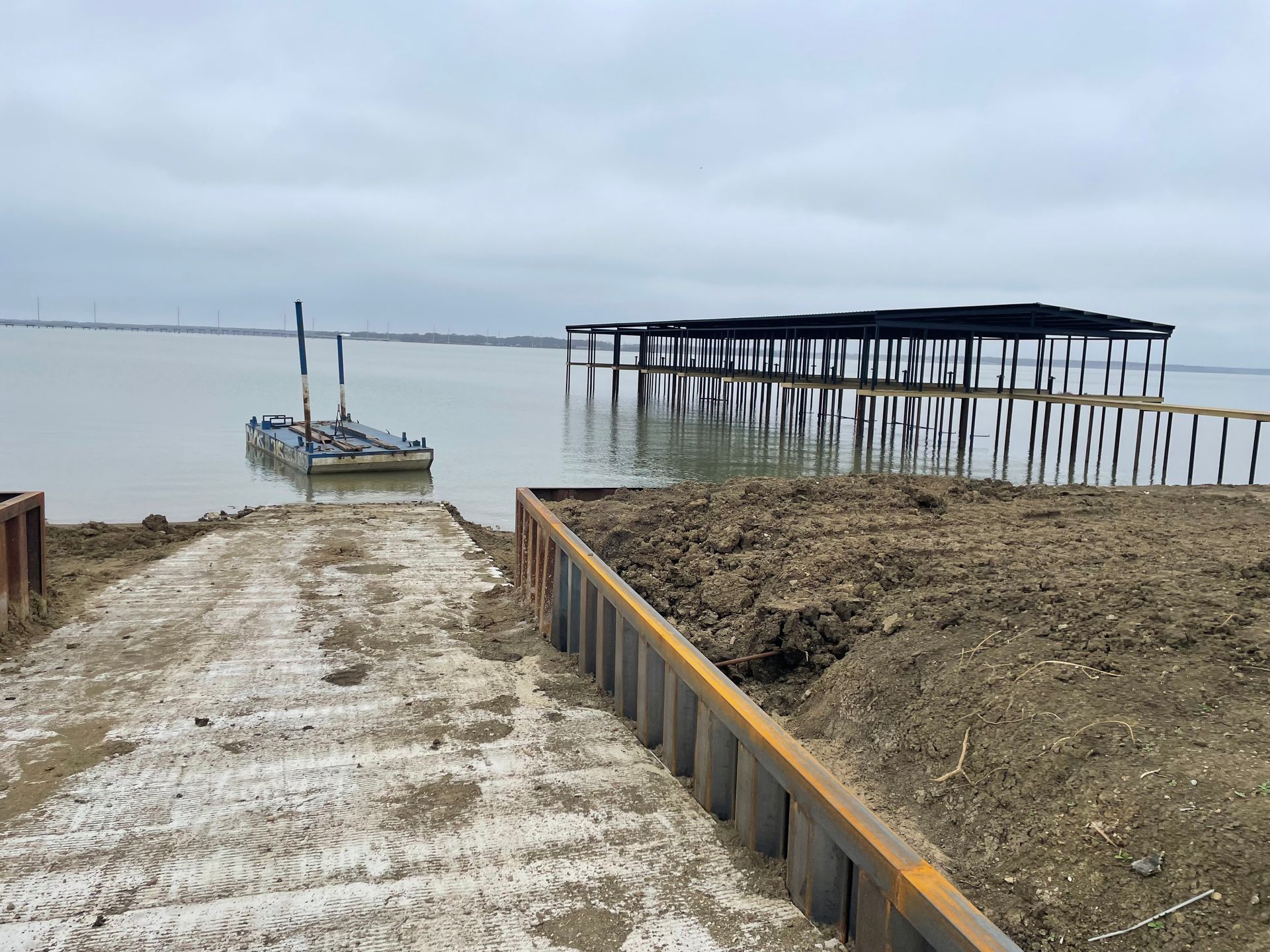 Small boat on a muddy boat ramp beside calm water and a weathered pier under an overcast sky