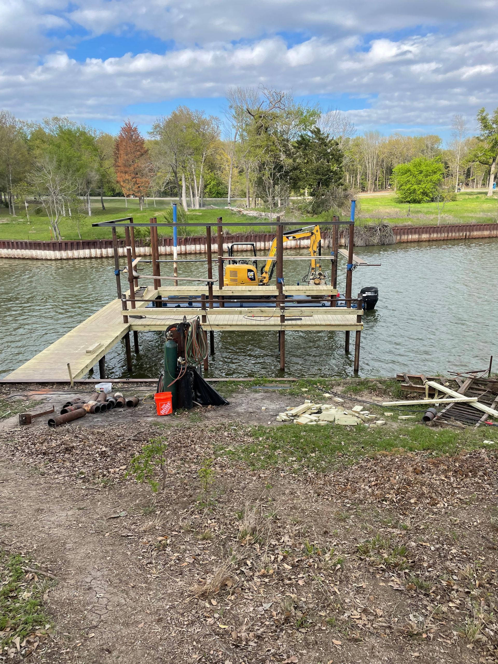 A dock on a pond with a yellow machine, framed by posts and surrounded by trees and grass.