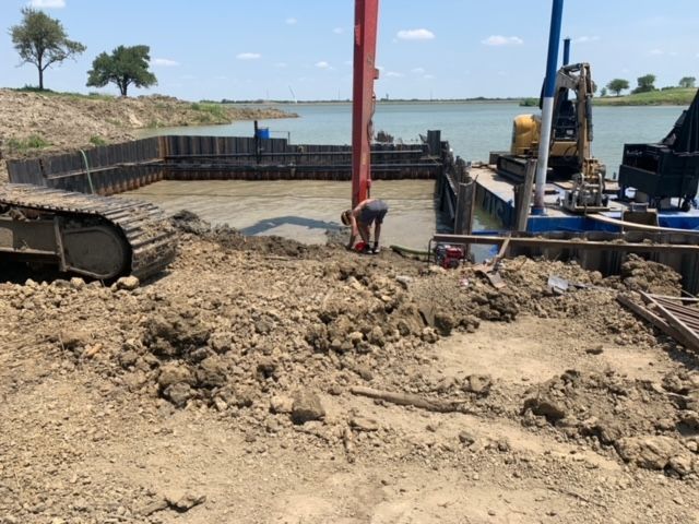 Construction site by water with excavator, red crane, and workers on muddy ground