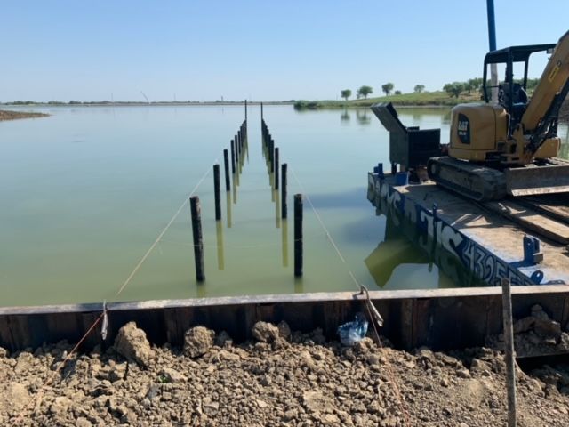 Calm water beside a construction site with posts in the water and an excavator on a platform.