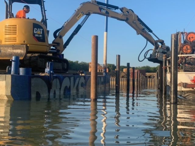Construction excavator working beside a flooded dock with metal posts and reflected water
