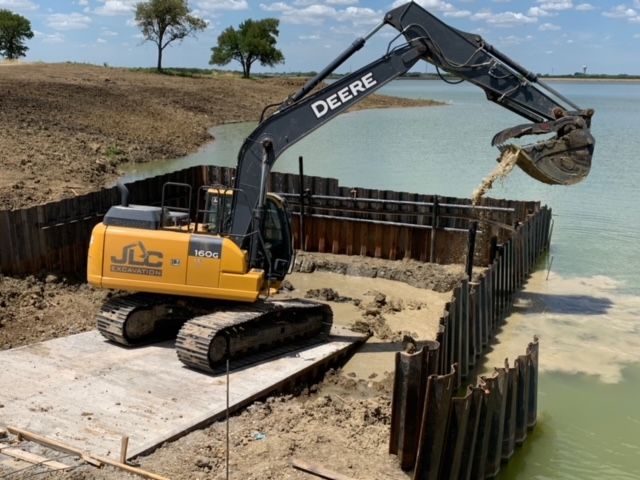 Yellow John Deere excavator dumping sand into a steel cofferdam beside water on a construction site
