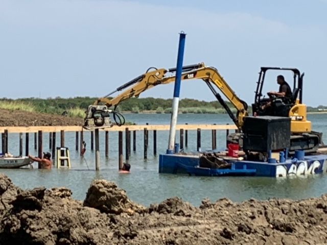 Yellow excavator on a floating platform driving piles in shallow water at a construction site