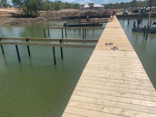 Wooden dock over green water leading to a shore with buildings and people in the distance