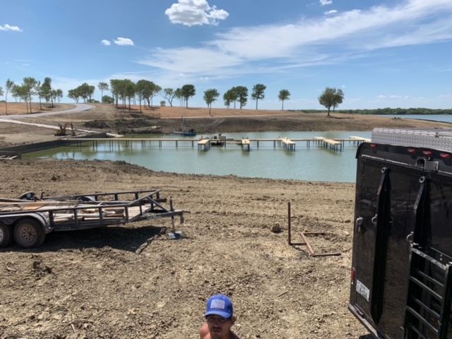 Dry, muddy pond with a small dock, trailers in the foreground, and sparse trees under a blue sky.