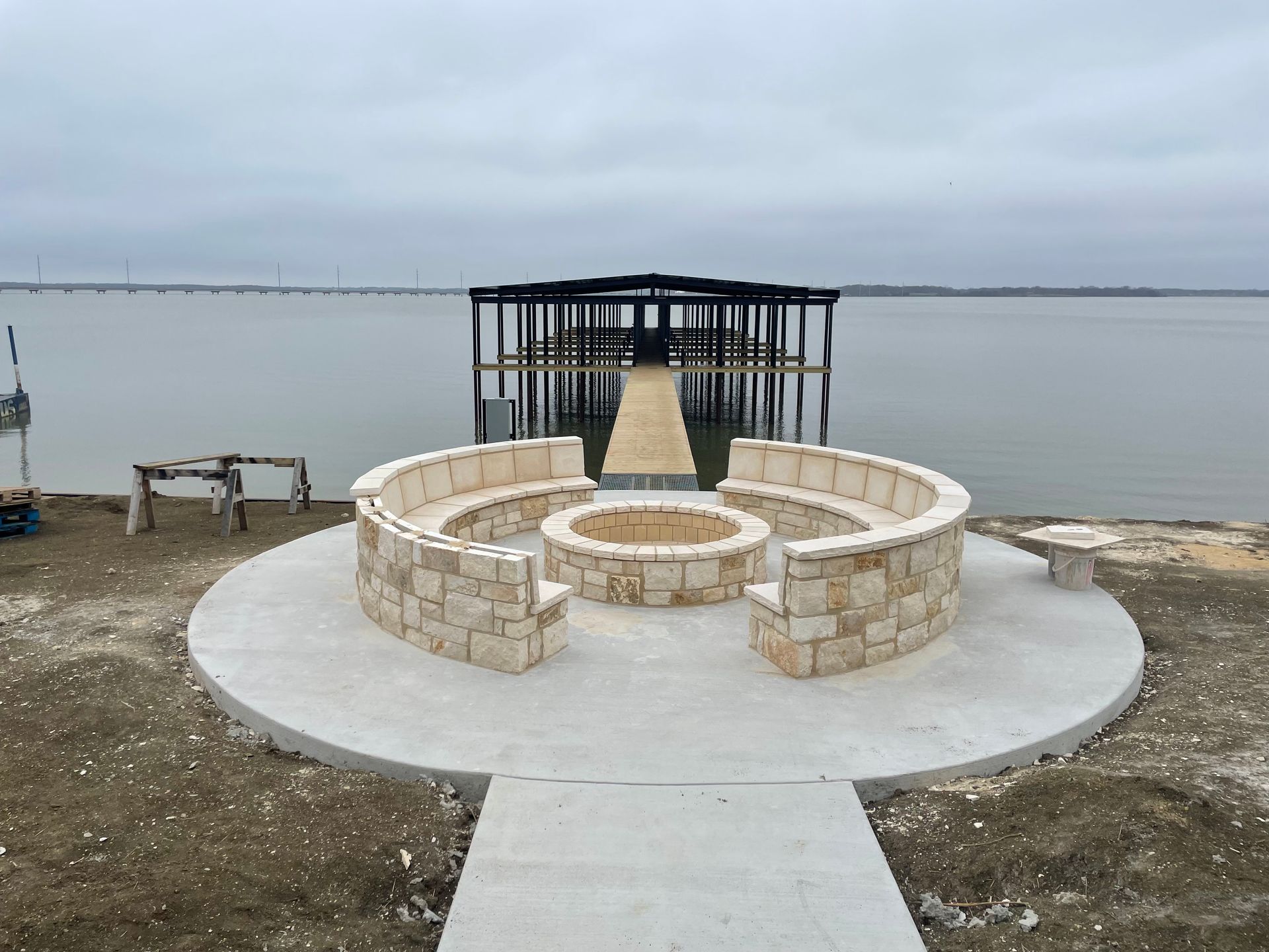 Stone circular seating area by a calm waterfront, with a central fire pit and dock behind it.