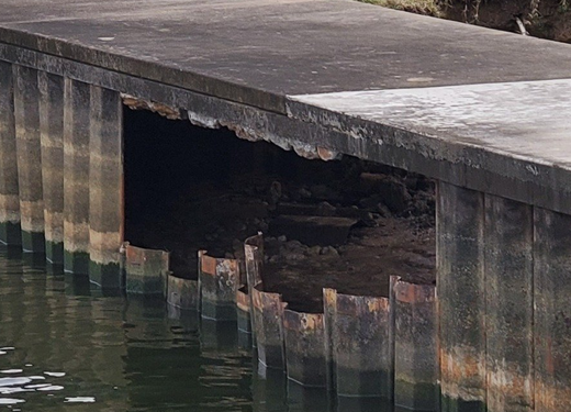 Damaged concrete seawall with a large hole above dark water, exposing erosion underneath