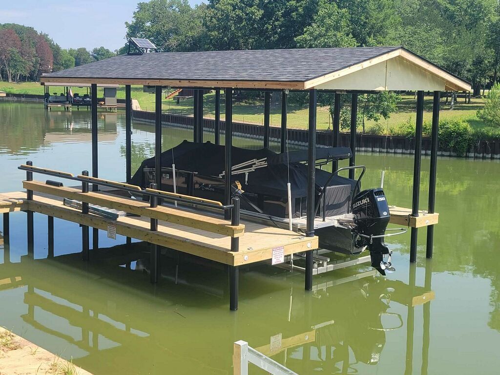 Covered boat dock on a calm lake with a black boat and wooden railings under a metal roof.