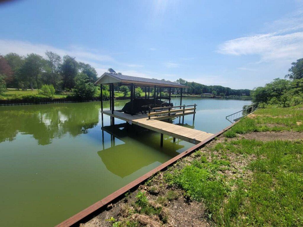 Covered dock on a calm green lake beside a grassy shore under a clear blue sky