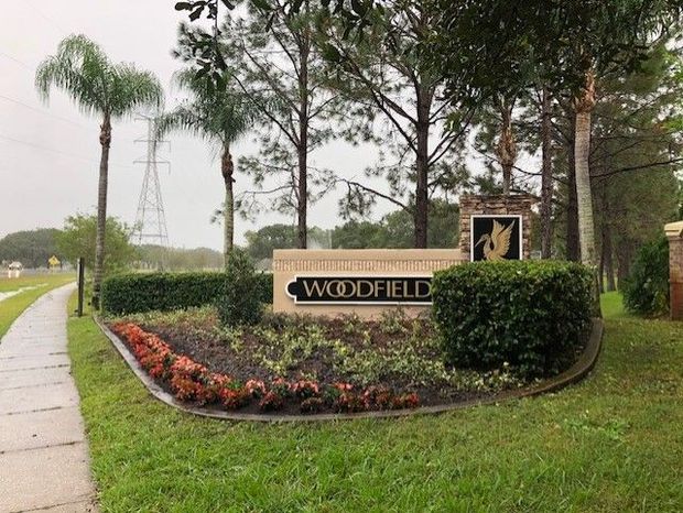 Woodfield neighborhood entrance sign beside a wet sidewalk, landscaped with flowers, hedges, and palm trees