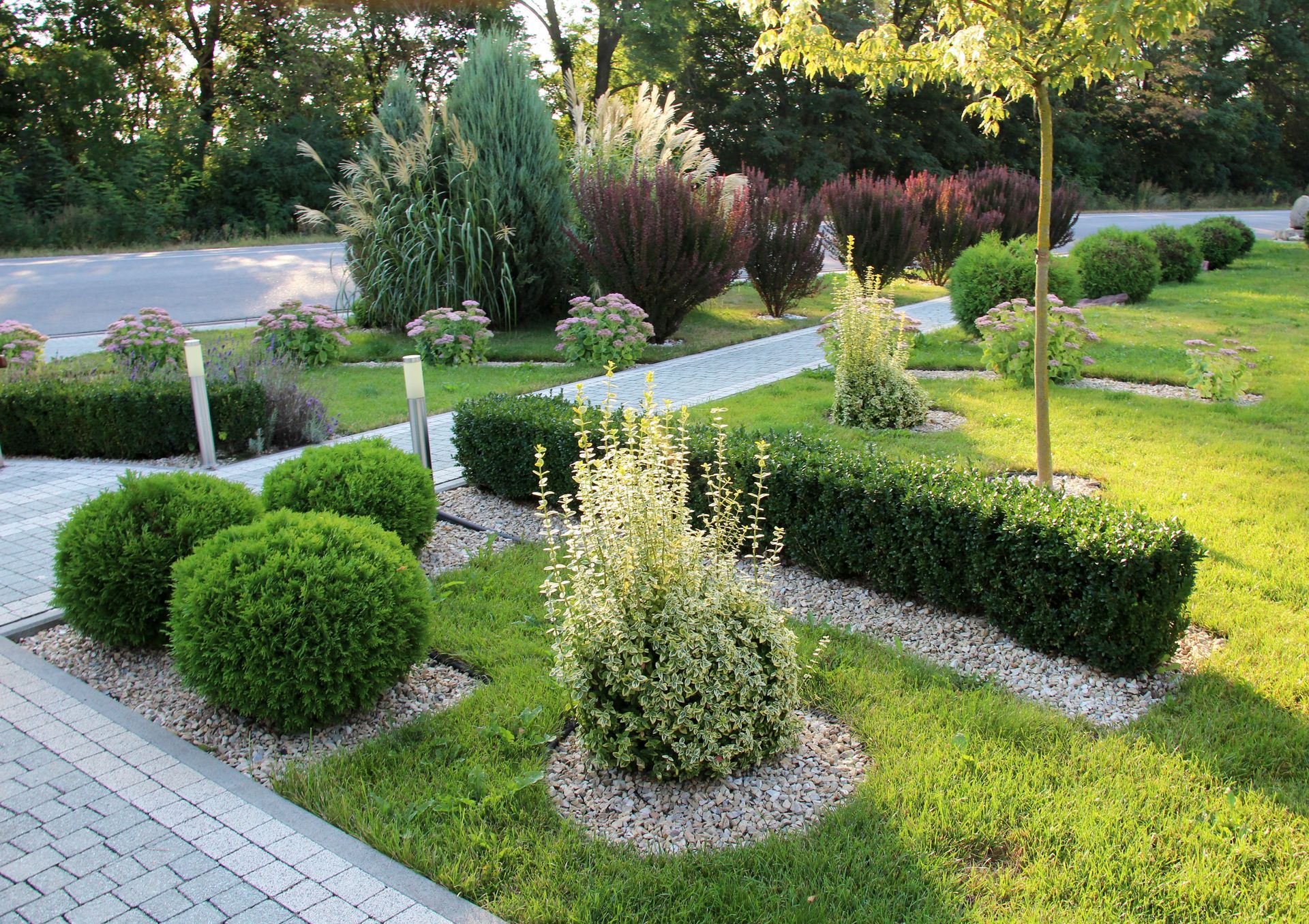 Manicured garden with shrubs, small trees, and white-edged flower beds beside a paved path Manicured garden with shrubs, small trees, and white-edged flower beds beside a paved path