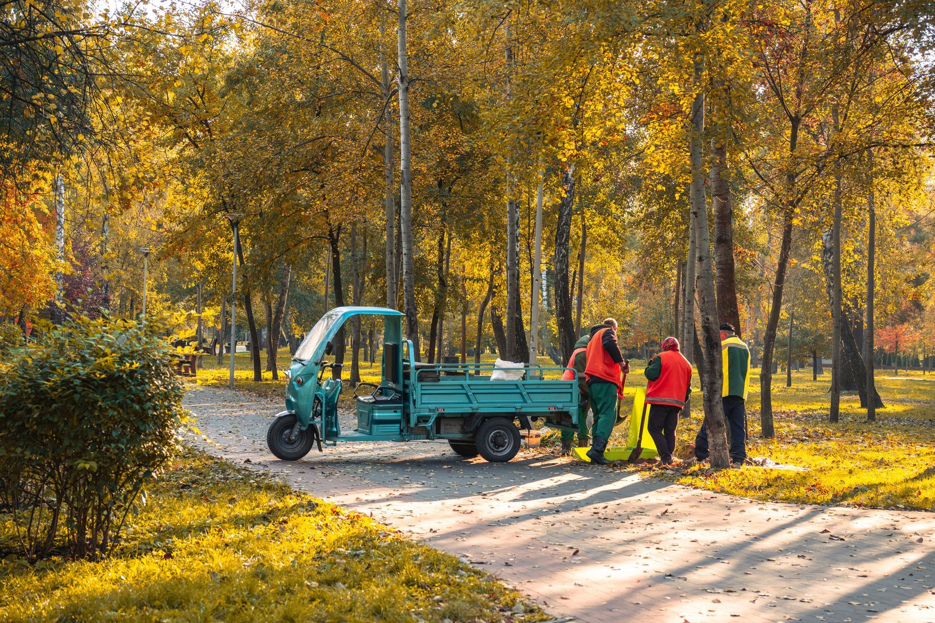 Workers in orange vests load debris into a teal utility truck in a park with autumn trees. Workers in orange vests load debris into a teal utility truck in a park with autumn trees.