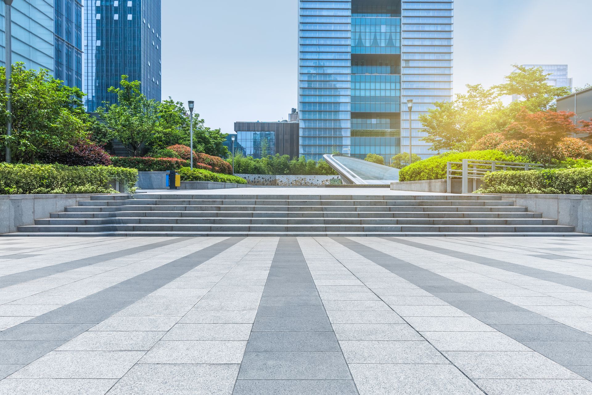 Modern plaza with gray tiled steps, green shrubs, and glass office towers in sunlight Modern plaza with gray tiled steps, green shrubs, and glass office towers in sunlight