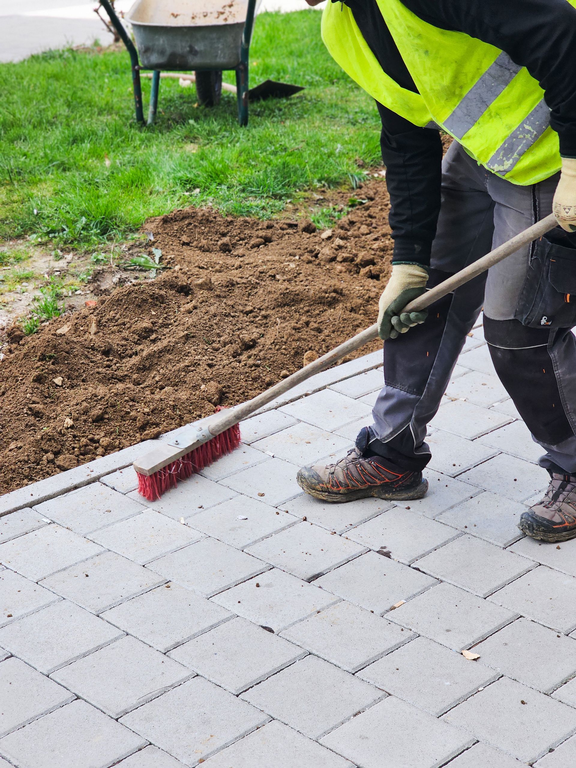 Worker sweeping dirt from a paved walkway with a red broom near a grass edge Worker sweeping dirt from a paved walkway with a red broom near a grass edge