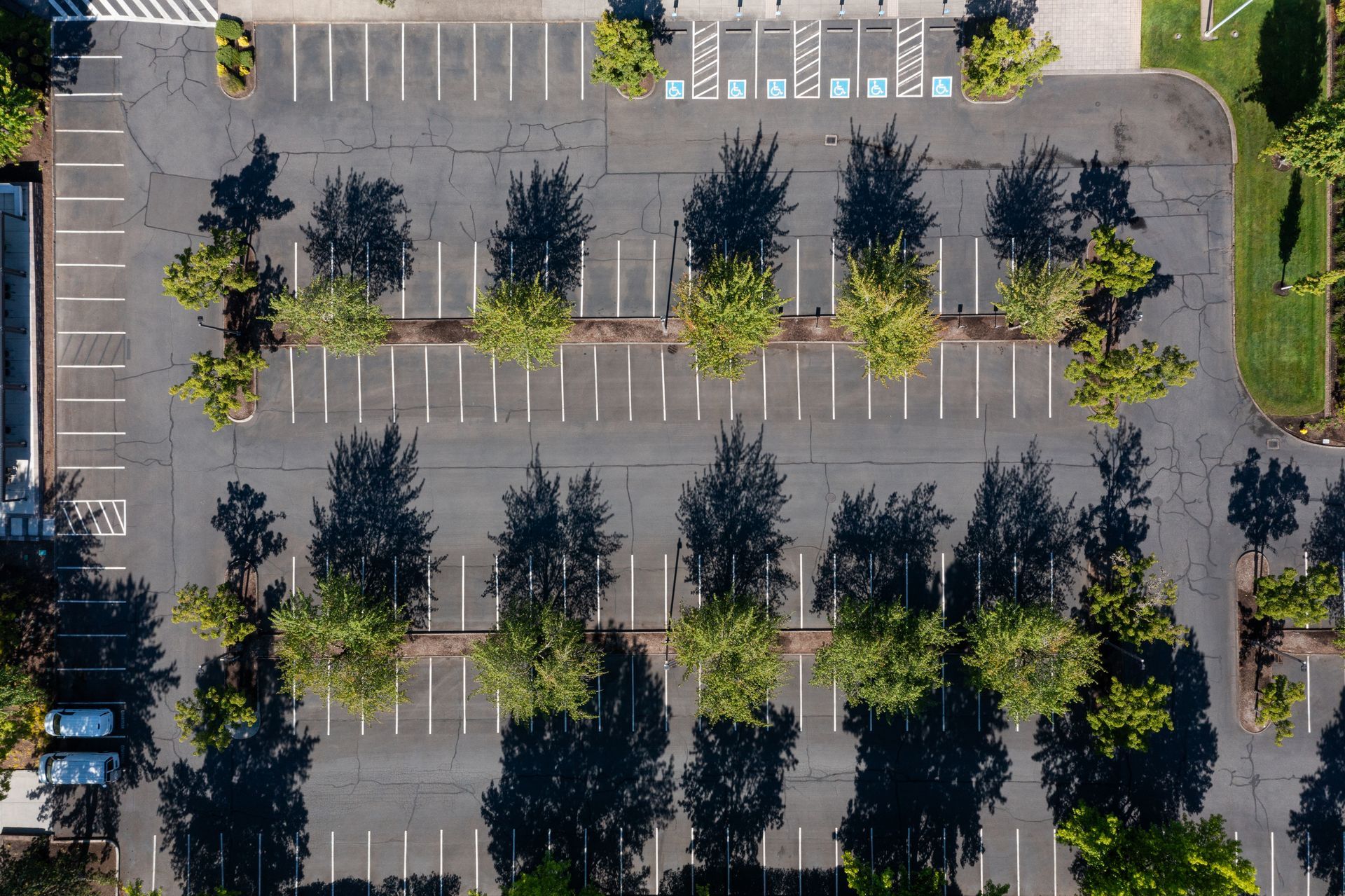 Aerial view of a mostly empty parking lot with trees casting shadows and a few parked cars. Aerial view of a mostly empty parking lot with trees casting shadows and a few parked cars.