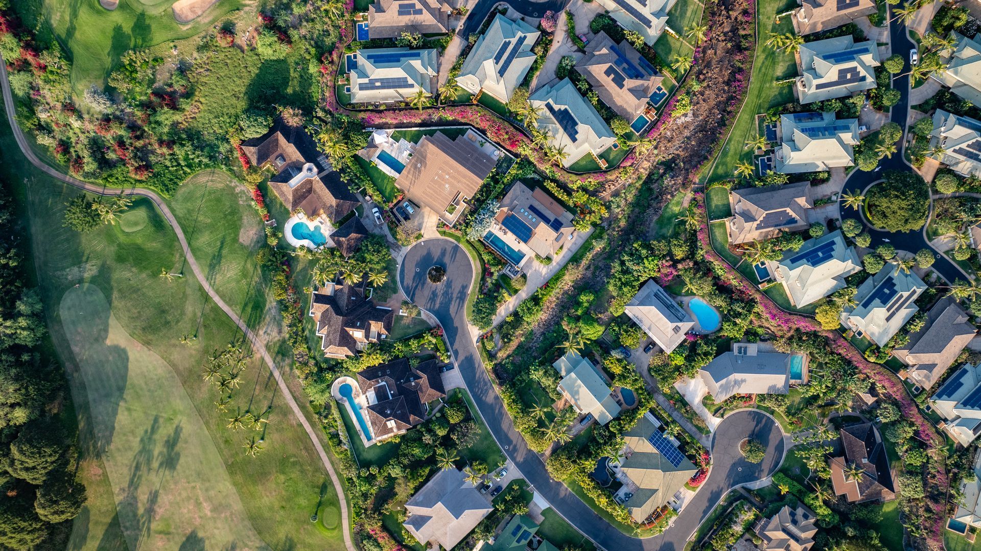 Aerial view of a suburban neighborhood with houses, roads, trees, and a swimming pool beside a green field Aerial view of a suburban neighborhood with houses, roads, trees, and a swimming pool beside a green field