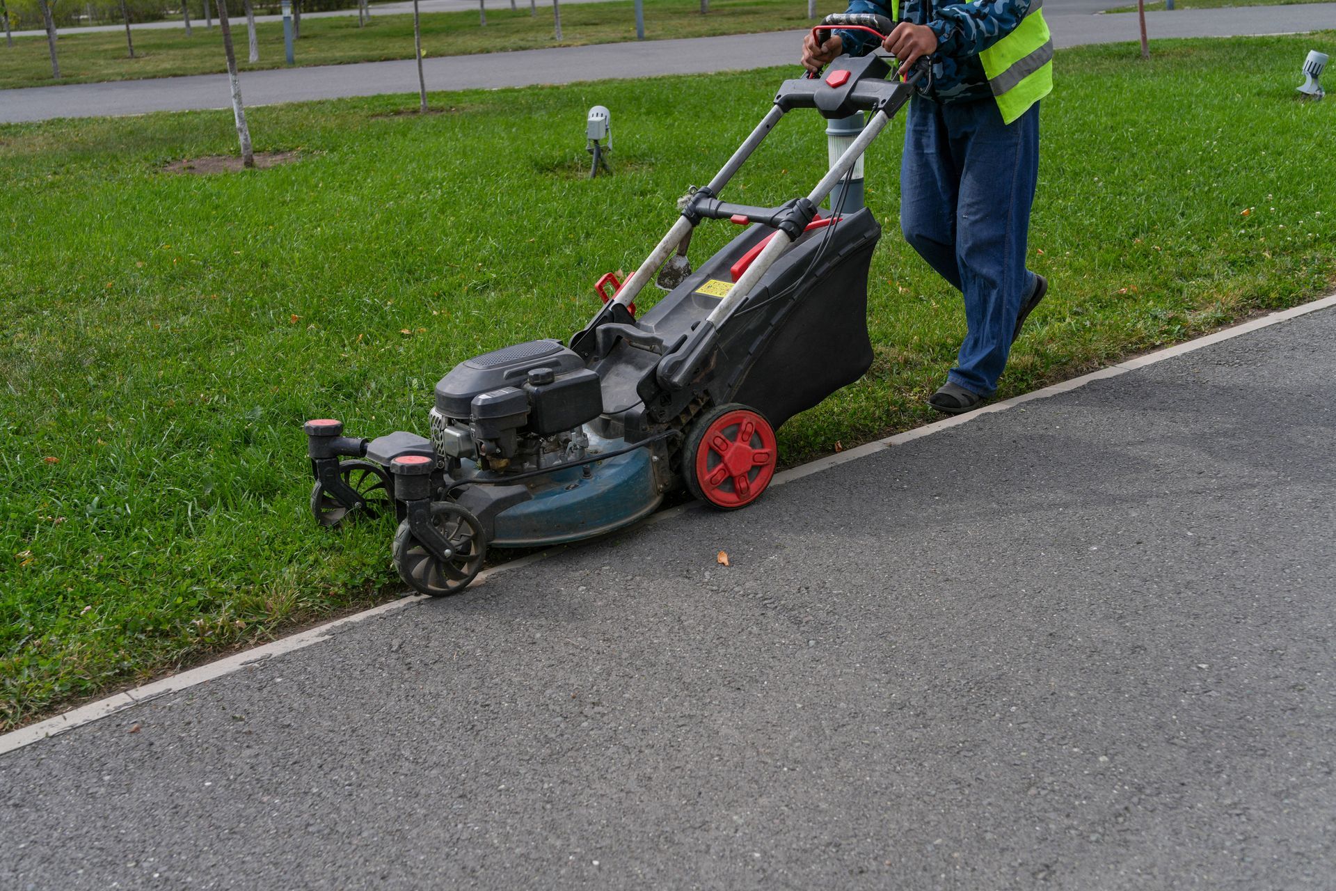 Person pushes a lawn mower along a roadside verge beside a paved street. Person pushes a lawn mower along a roadside verge beside a paved street.