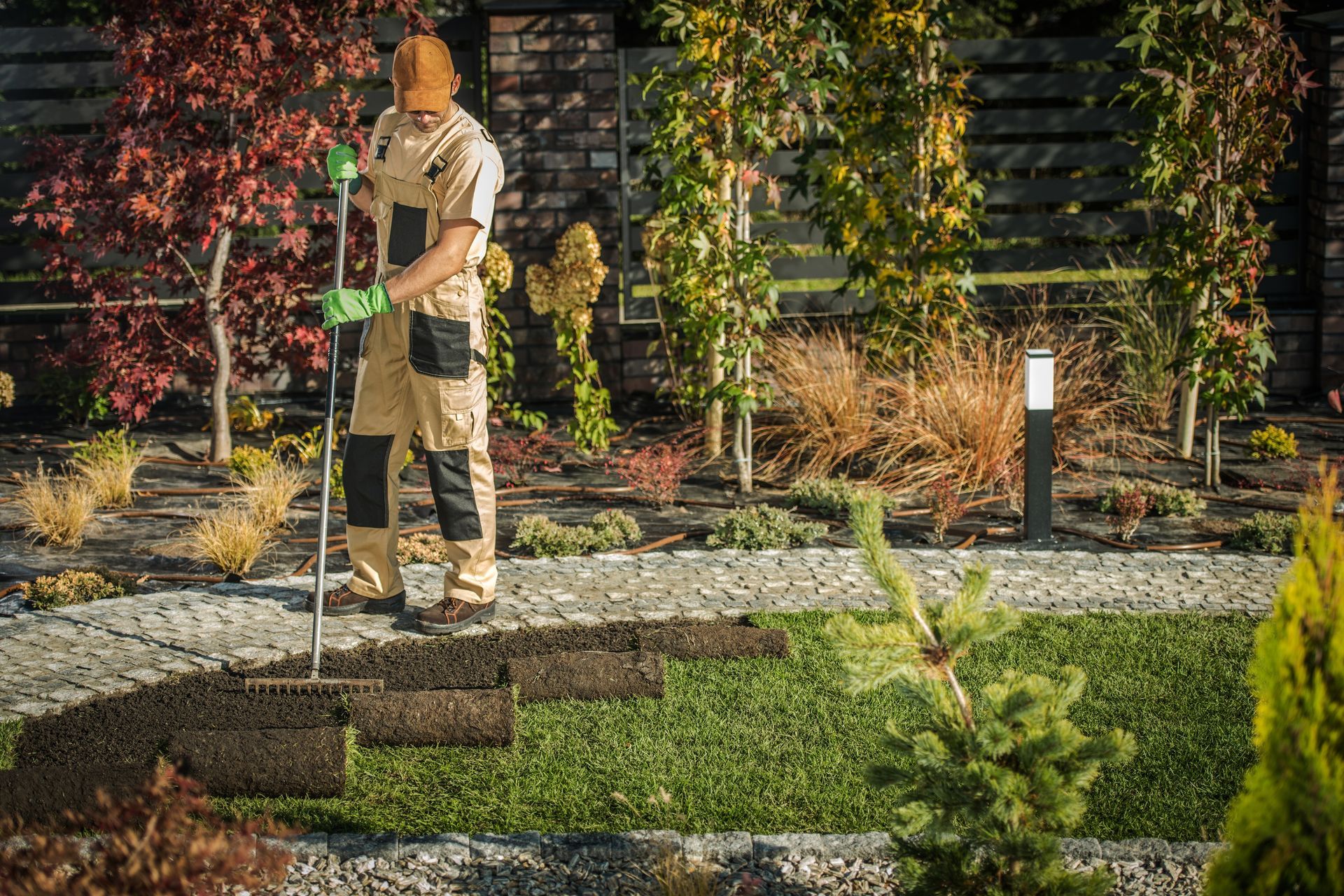 Person in a tan outfit stands on a walkway in a landscaped garden, holding a shovel.