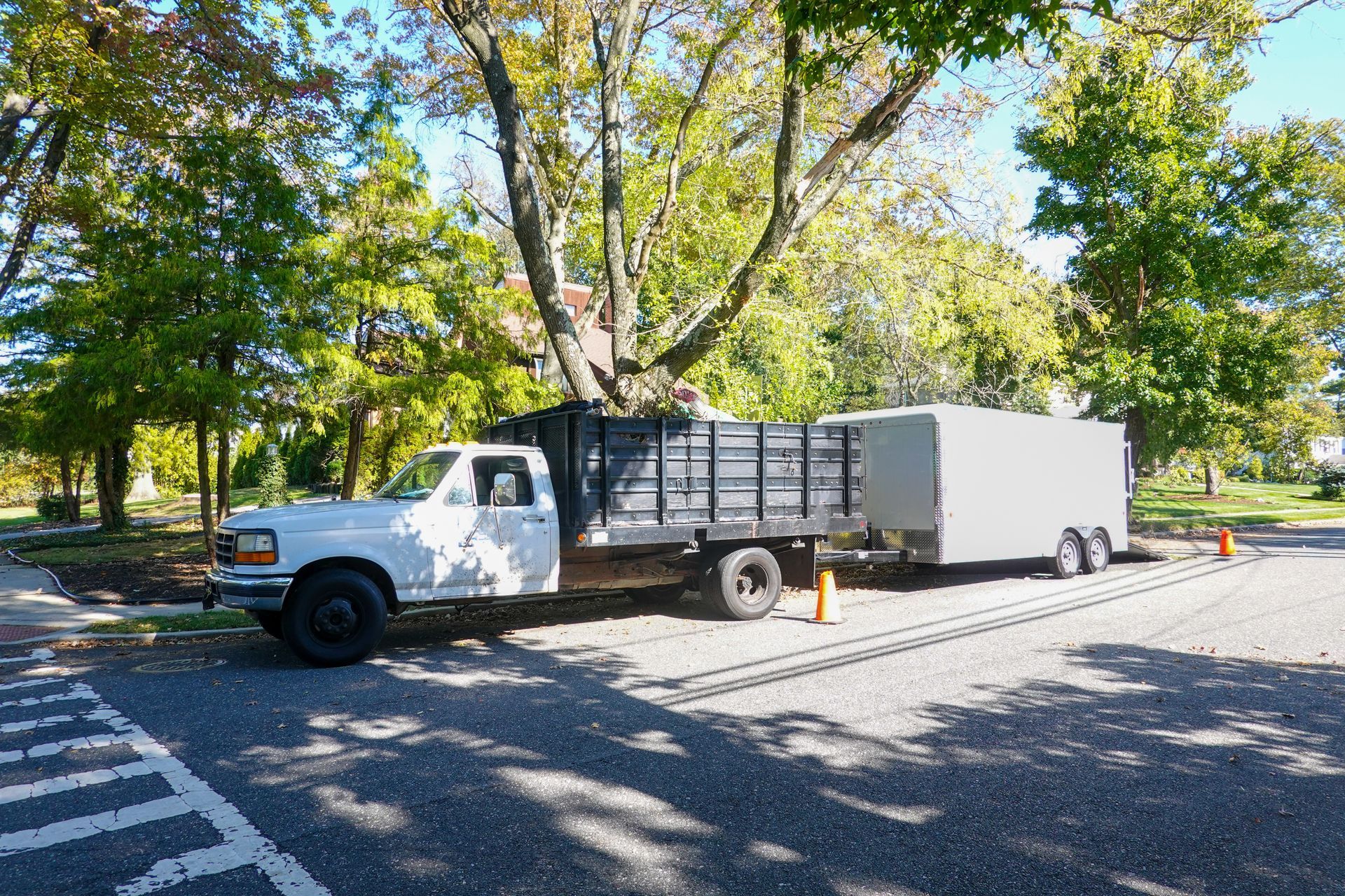 White utility truck parked beside a gray enclosed trailer under trees on a sunny street White utility truck parked beside a gray enclosed trailer under trees on a sunny street