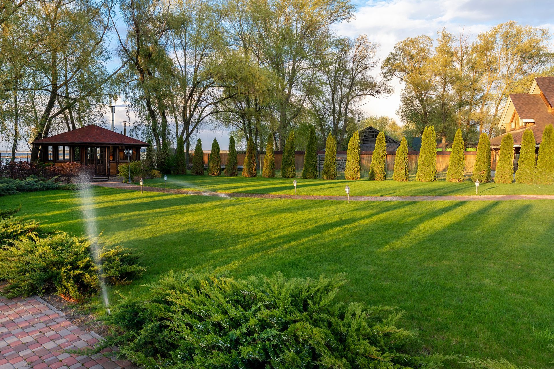 Sunlit landscaped garden with green lawn, trees, shrubs, and a gazebo by a brick path Sunlit landscaped garden with green lawn, trees, shrubs, and a gazebo by a brick path