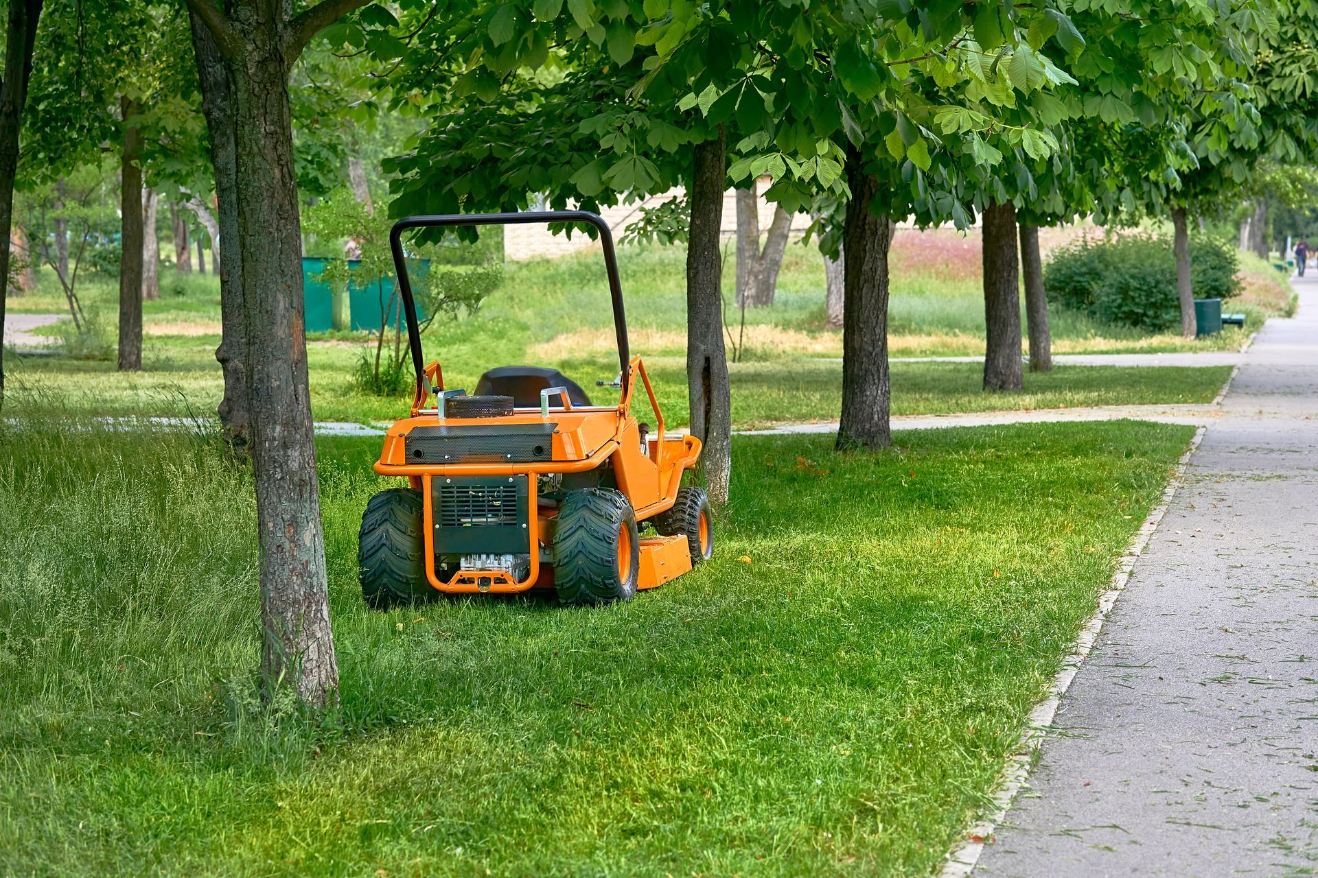 Orange lawn mower parked on grass beside a tree-lined path in a park. Orange lawn mower parked on grass beside a tree-lined path in a park.
