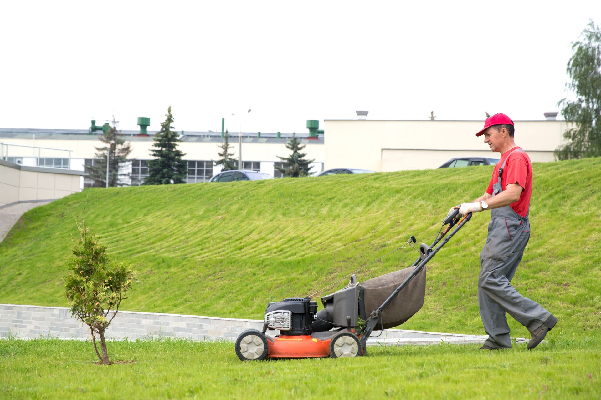 Person pushing a red lawn mower on a grassy hill near a building