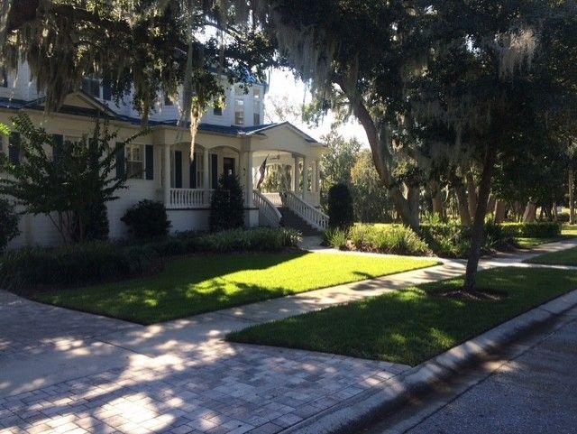 Tree-shaded suburban house with a porch, bright green lawn, and sunlit sidewalk by the street