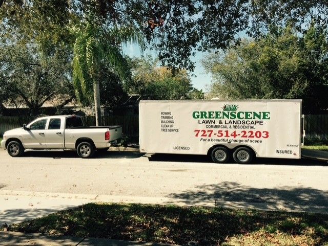 White pickup truck towing a Greenscene trailer parked under trees on a sunny street