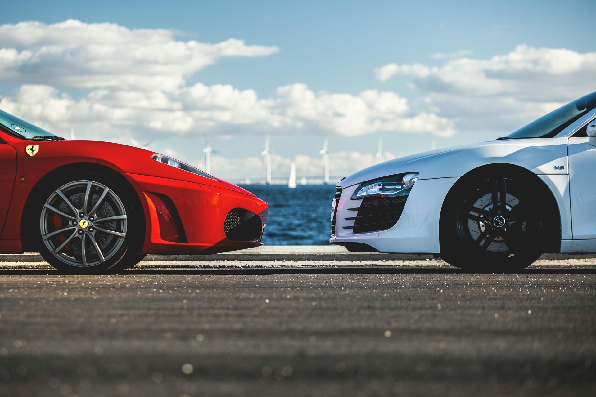 Une Ferrari rouge et une Audi blanche, voitures de sport garées côte à côte, près d'un point d'eau avec des moulins à vent et un ciel bleu.