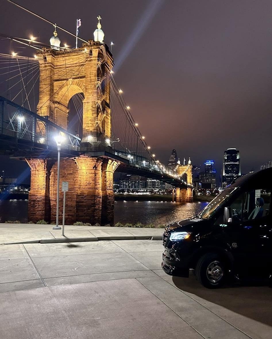 Night view of a lit suspension bridge over water, with a black van parked in the foreground.