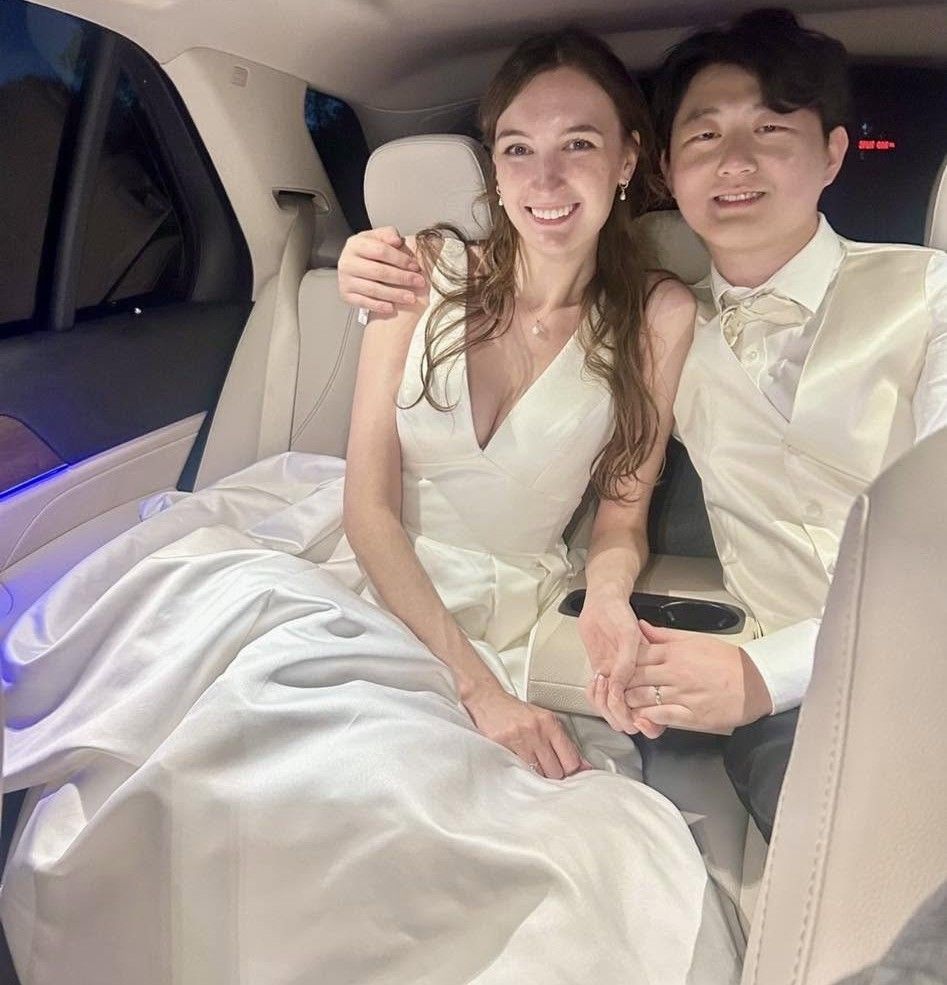 Two people in formal white attire smiling together in the back seat of a car