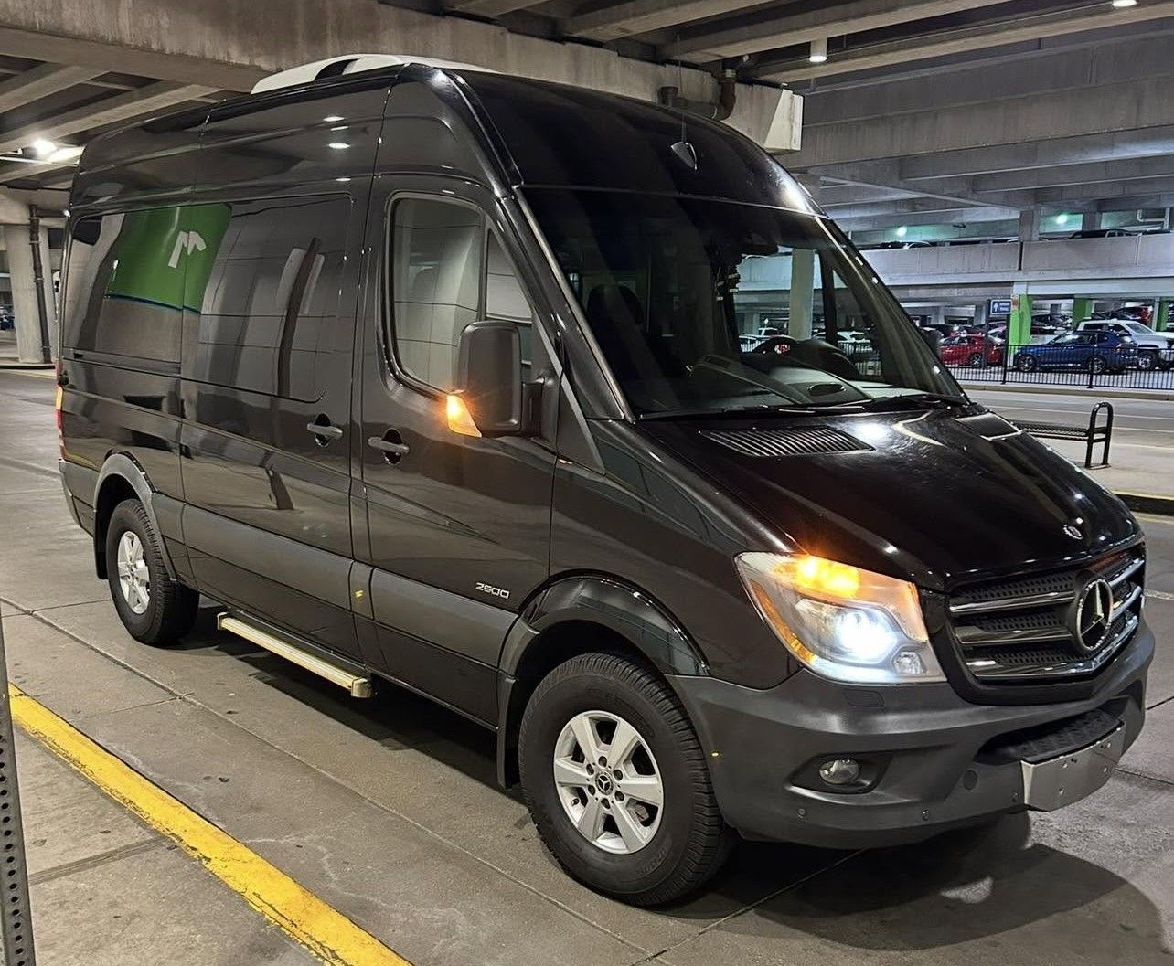 Dark gray Mercedes van parked in a covered garage, front-left view with headlights on