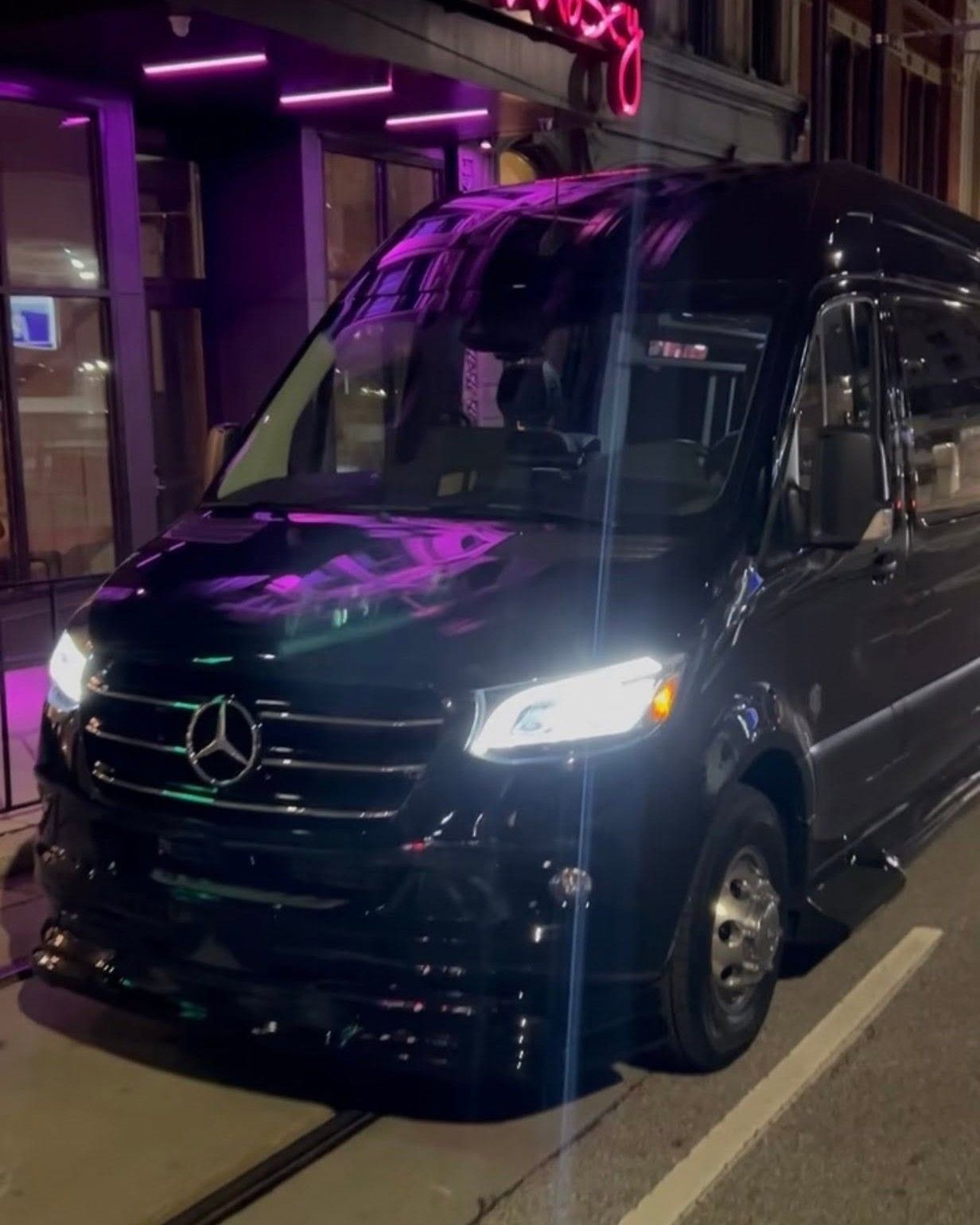 Black Mercedes van parked at night on a city street, lit by purple neon reflections.
