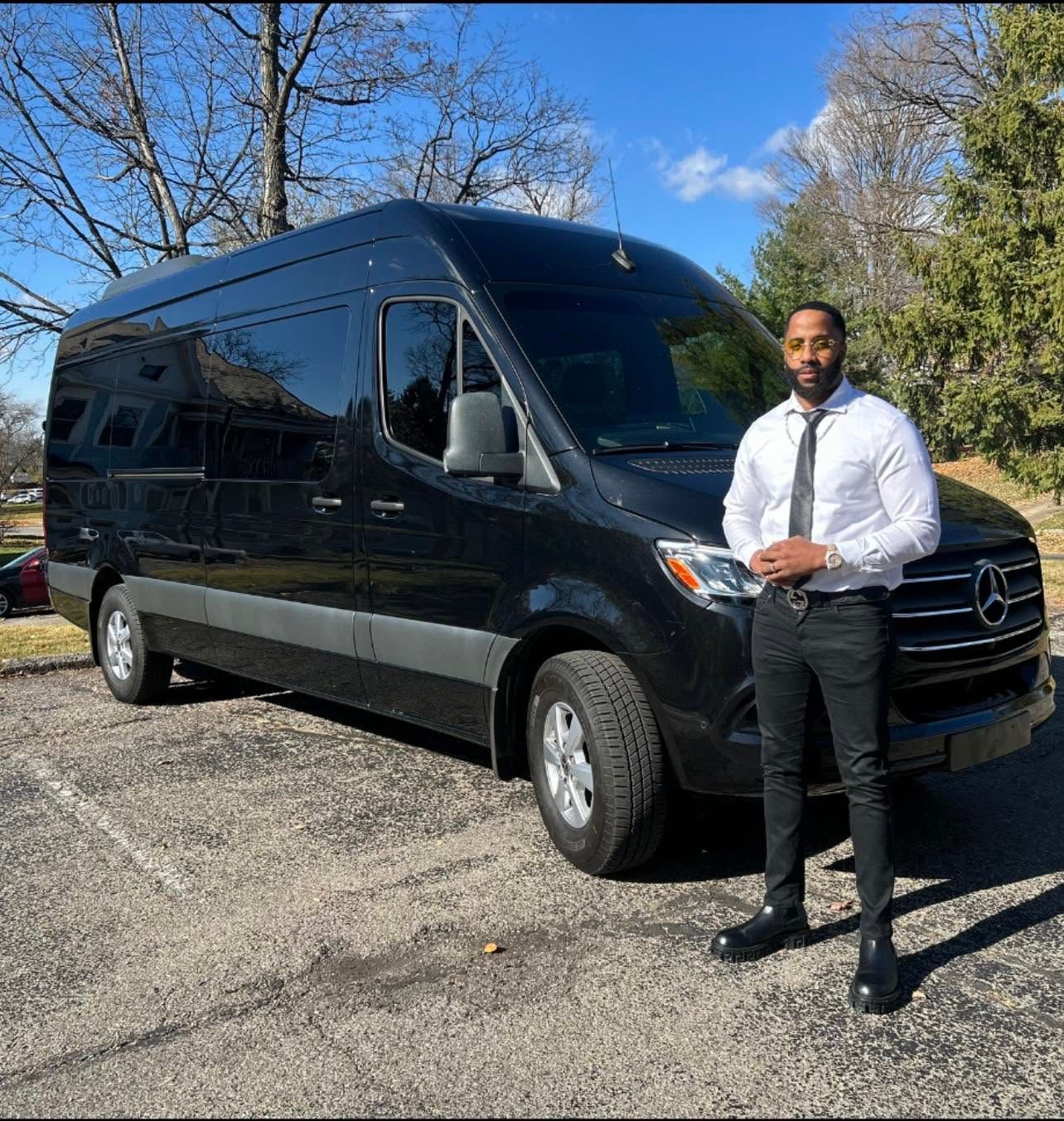 Man standing beside a black passenger van on a sunny gravel lot with trees in the background