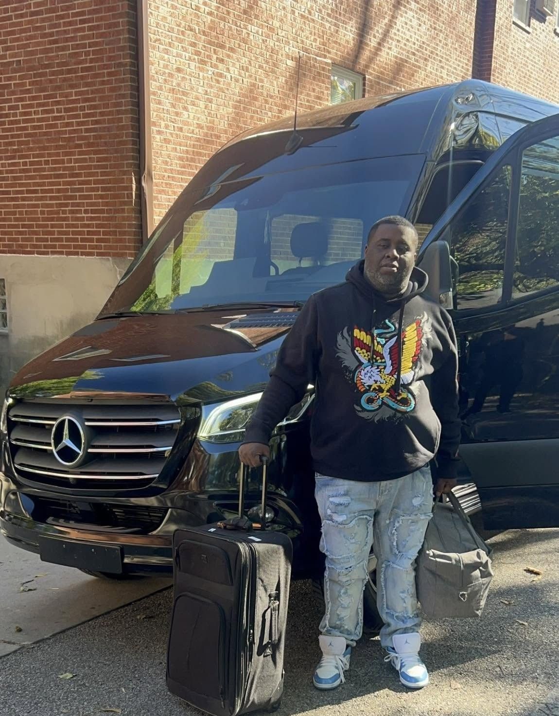 Person standing beside a black Mercedes van with a suitcase, outdoors by a brick building.