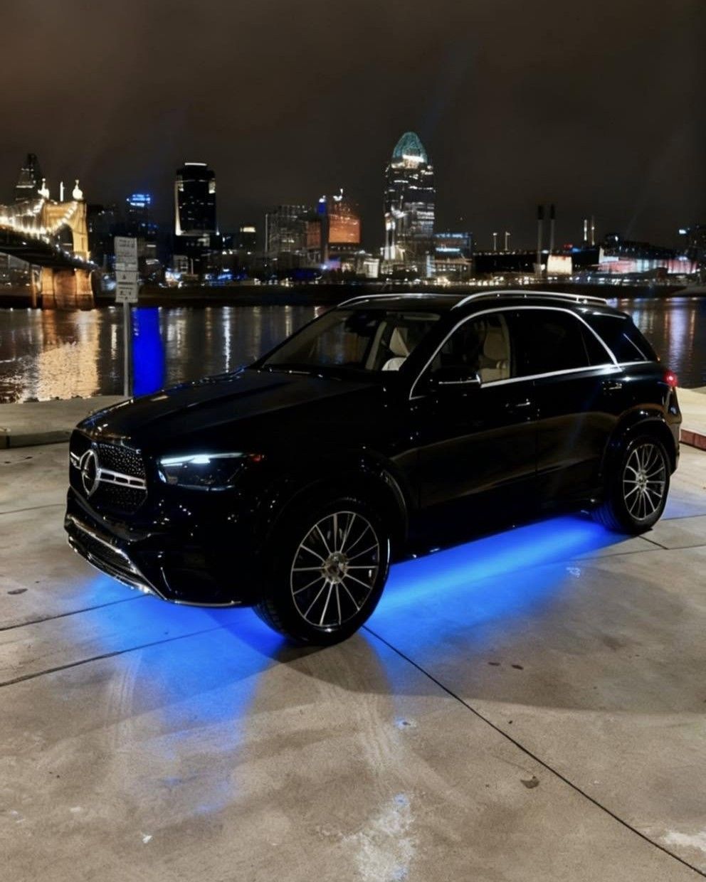 Black SUV with blue underglow parked on a waterfront at night, city skyline in the background.