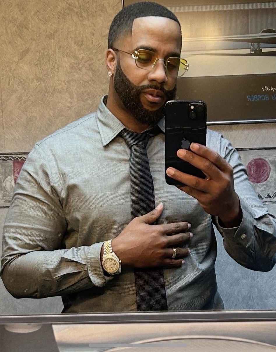 Mirror selfie of a man in a gray shirt and tie, wearing gold glasses and a watch.
