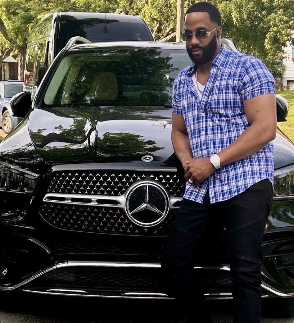 Man leaning against a black Mercedes SUV in a leafy driveway, wearing a blue plaid shirt and sunglasses