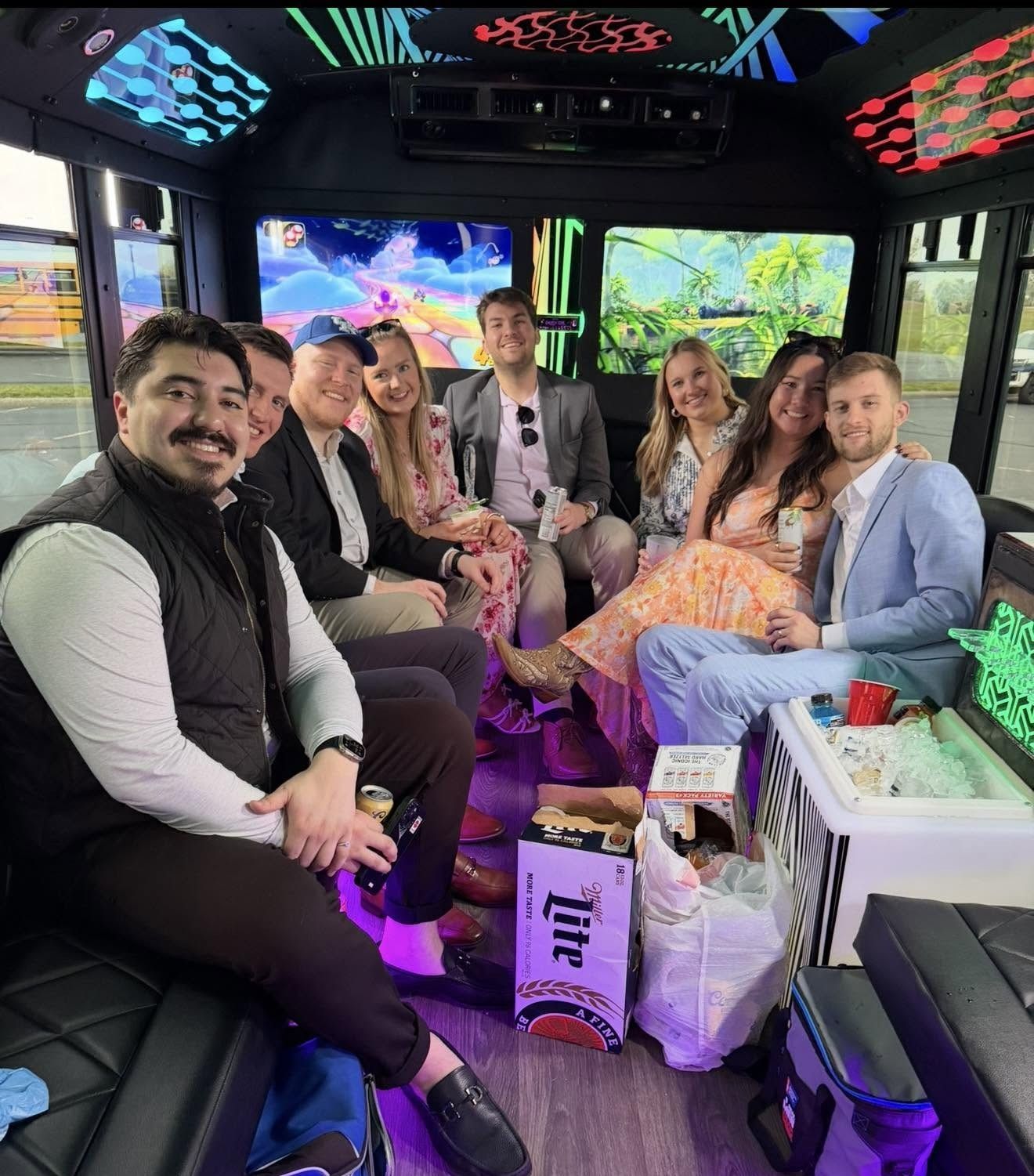 Group of smiling people seated inside a neon-lit party bus with drinks and coolers.